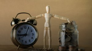 a wooden figure posing one hand on a clock and the other on a money jar